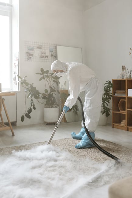 A person using a handheld vacuum cleaner with a black body and a flexible hose, cleaning a traditional patterned area rug with floral motifs in muted tones of beige, green, and orange. The vacuum atta