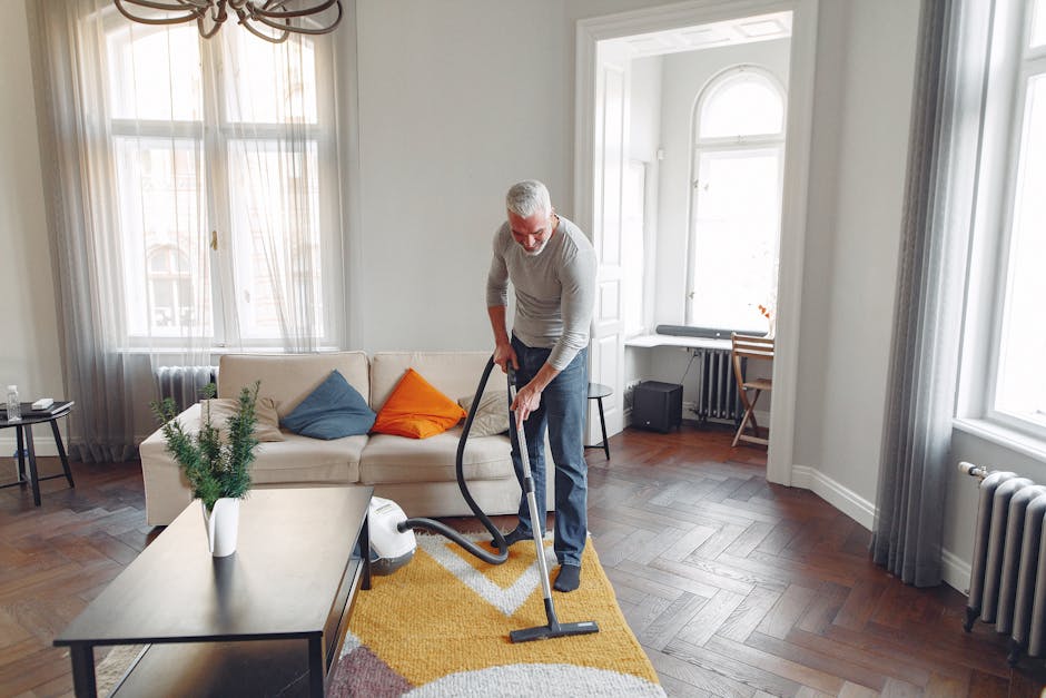 A man with grey hair and a beard is using a vacuum cleaner to deep clean a living room with wooden parquet flooring, a beige sofa adorned with blue and orange cushions, a coffee table with a potted pl
